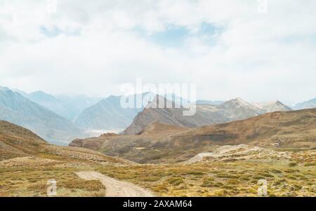 Strada che collega villaggi fiancheggiati da montagne dell'Himalaya sotto overcast, cielo e arbusti in primo piano vicino a Tashigong, Himachal Pradesh, India. Foto Stock