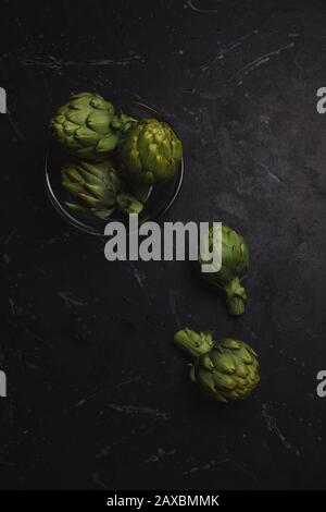 Carciofi in una ciotola di vetro e più verdure sul tavolo, su uno sfondo scuro afflitto Foto Stock