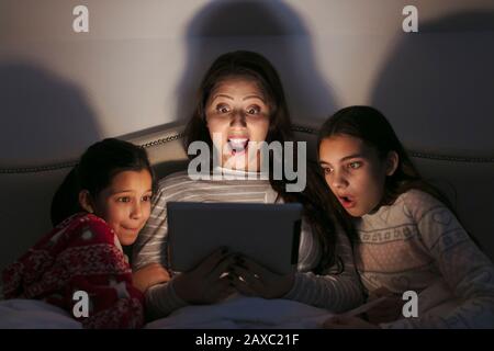 Madre e figlie sorpresi nel guardare film su un tablet digitale in camera oscura Foto Stock