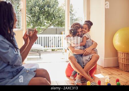 Madre con macchina fotografica telefono fotografare padre e figlie abbracciando Foto Stock