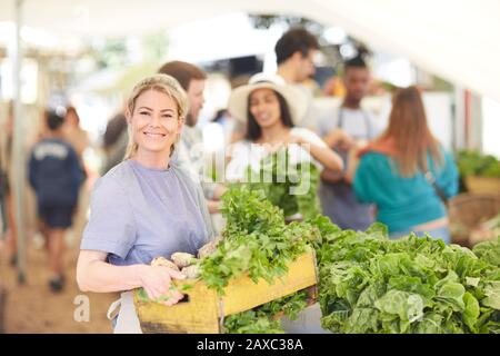 Ritratto donna sorridente e fiduciosa che lavora, portando crate di verdure al mercato contadino Foto Stock