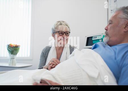 Una donna anziana che si prende cura di visitare, un marito confortante che riposa nel letto dell'ospedale Foto Stock