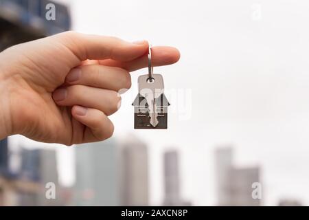 La mano che tiene una chiave ad una casa di sogno contro lo sfondo dei lucernari della città. Il concetto di locazione o vendita di una casa o appartamento, ipoteca e re Foto Stock