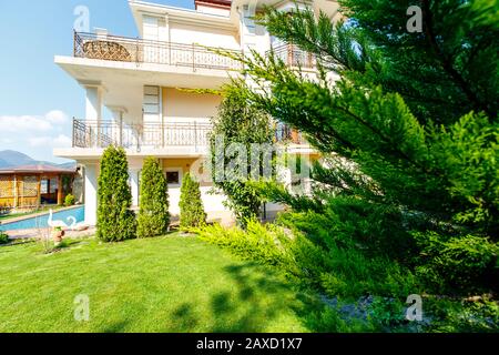 Il cortile di un classico cottage bianco a tre piani con ringhiere e colonne in ferro battuto. Molla. Di fronte al cottage c'è un prato verde, tre Foto Stock