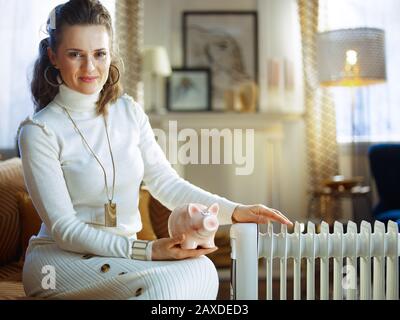 donna moderna e sorridente in maglione bianco e gonna in casa moderna nella soleggiata giornata invernale seduto sul divano vicino bianco radiatore di olio elettrico tenendo il divieto piggy Foto Stock