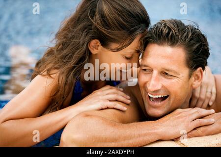 Una donna sorridente di media età che sussurra vicino ai suoi fidanzati orecchio mentre in acqua al bordo di una piscina. Foto Stock