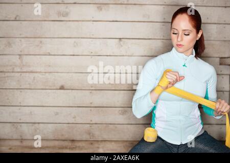 La giovane donna avvolge una mano con le mani di boxe gialle mentre si siede su una panca di legno davanti a una parete di legno. Foto Stock