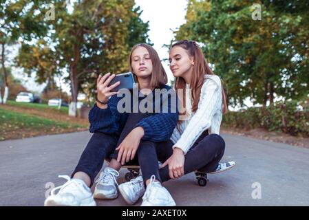 Due ragazze studentesse di 12-14 anni, in estate in un parco cittadino, guardare video su uno smartphone, sedersi su uno skateboard, abbigliamento casual, una pausa a scuola e. Foto Stock