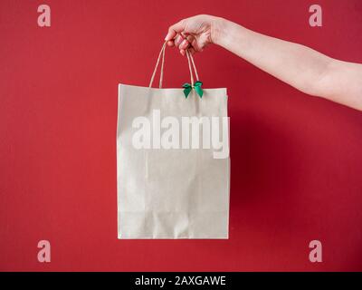 La mano della donna che tiene il sacchetto di carta riciclato bianco decora con l'arco verde piccolo su sfondo rosso della parete. Foto Stock