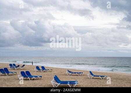 Tavira, Portogallo - 23 gennaio 2020: Un pescatore anziano getta la sua pole e la fila fuori durante i mari grezzi di inverno. Sedie da spiaggia blu vuote in primo piano Foto Stock