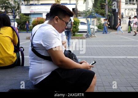 Antipolo City, Filippine - 11 febbraio 2020: L'uomo filippino si siede su una panchina del parco per rilassarsi e utilizzare il suo smartphone. Foto Stock