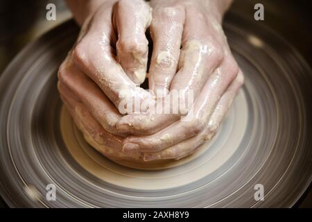 Il vasaio principale piegato insieme le mani sculpts un prodotto di creta su una ruota del vasaio primo piano. Pelle della mano macchiata con argilla e bagnata Foto Stock