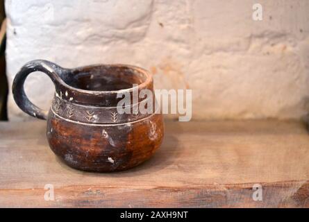 Vecchia tazza di argilla si erge su una superficie di legno vicino a una parete di mattoni bianchi Foto Stock