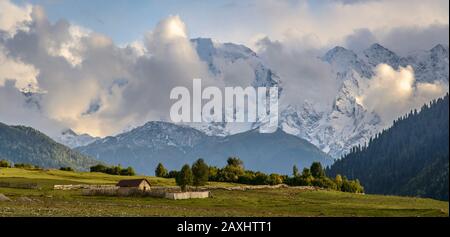 Piccola casa in pietra circondata da recinzione in legno sul prato alpino. Alte montagne coperte di neve e nuvole sullo sfondo. Foto Stock