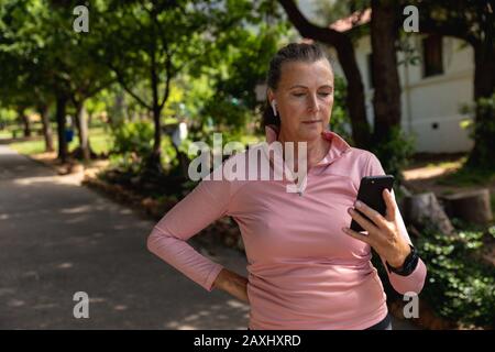 Vista frontale di una donna caucasica matura di mezza età che lavora nel parco, utilizzando smartphone e auricolari, preparandosi ad allenarsi Foto Stock