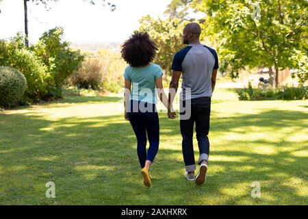 Weekend di divertimento a casa insieme. Vista posteriore di una coppia di gara mista che cammina nel giardino e tiene le mani durante una giornata di sole Foto Stock