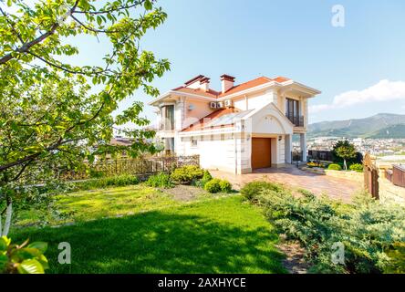 La facciata di un classico cottage mediterraneo a due piani in primavera. Il cottage è bianco con un tetto di tegole rosse. Di fronte al cottage c'è un Foto Stock