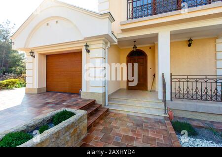 La facciata di un cottage classico bianco a due piani con giardino frontale. Porta d'ingresso scolpita, cancello auto, piastrelle beige, alberi. Foto Stock