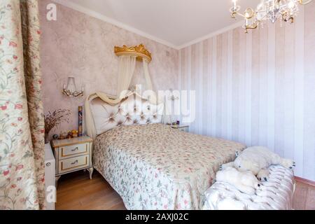 Elegante camera da letto Royal bianca con un grande letto matrimoniale. Retro bianco Intagliato con un piccolo baldacchino, comodini bianchi con gambe curve. Parete a righe chiare Foto Stock