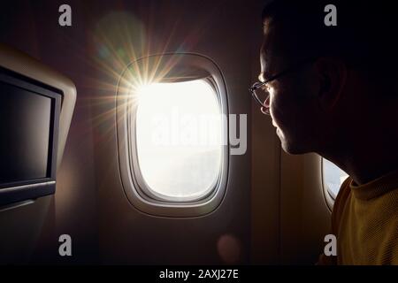 Giovane uomo con occhiali che guarda attraverso la finestra dell'aeroplano durante la giornata di sole. Foto Stock