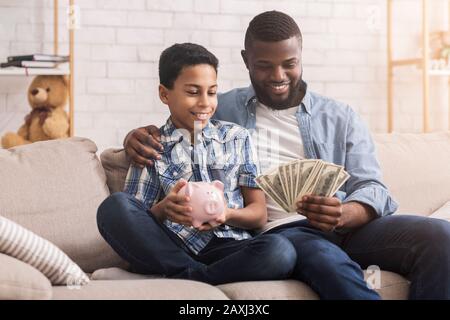 Risparmi Personali. Piccola Ragazza Afro Mettendo Soldi A Piggybank, Padre Che Lo Tiene Foto Stock