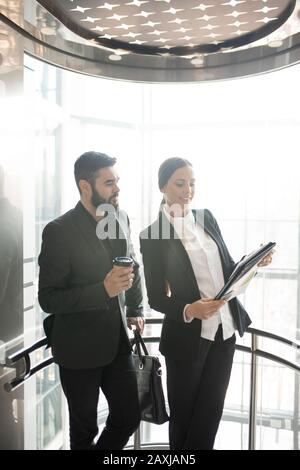 Sorridente giovane donna d'affari in tuta mostrando documenti di lavoro al collega mentre si spostano in ascensore Foto Stock
