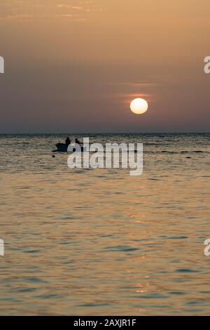 Concentrati sulla navigazione in barca con silhouette di due uomini. Cerchio di sole in spiaggia al tramonto di fronte all'acqua dorata del Mar Ionio con nuvole rovesate. Crepuscolo come visto da Ksamil, Albania, primavera sera Foto Stock