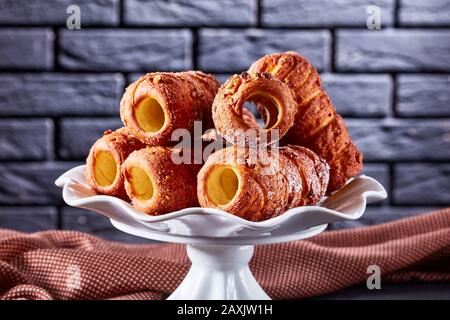 Torta al camino ungherese o kurtos kalacs - dolce dolce laminato servito su un piatto bianco su uno sfondo di cemento scuro di fronte a un bric nero Foto Stock