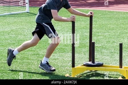 Un ragazzo di scuola superiore spinge una slitta gialla con un peso di 25 libbre su di essa durante la pratica del circuito. Foto Stock