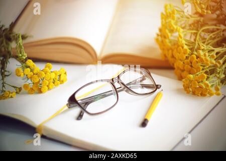 Sul posto di lavoro, un libro per la lettura, un notebook per la scrittura, una matita, occhiali e bouquet di colore giallo brillante, illuminato dalla luce del sole, sono su t. Foto Stock