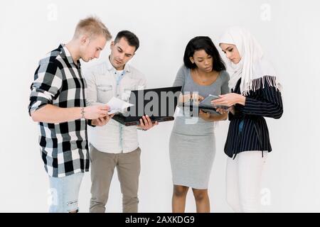 Gruppo Di Quattro Studenti Multietnici, che lavorano insieme, utilizzando notebook e tablet, in piedi Su sfondo bianco. Commercio internazionale Foto Stock