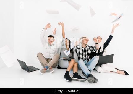 Vincitori aziendali, esame finale superato. Gruppo multirazziale di giovani allegri in abbigliamento casual seduto sul pavimento, mantenendo le braccia sollevate e. Foto Stock