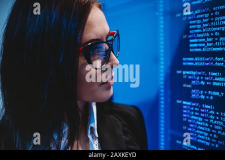 Donna attraente con occhiali che guardano i codici sullo schermo in ufficio da vicino Foto Stock