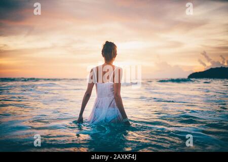 Vista posteriore della giovane donna indossando un abito bianco a piedi nell'oceano al tramonto. Foto Stock