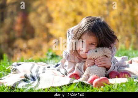 Ragazza allegra che abbraccia un coniglio giocattolo, mele rosse sdraiate sull'erba durante il picnic nella soleggiata giornata autunnale nel parco Foto Stock