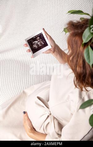 Ritratto di bella signora curly incinta in abito beige che tiene ecografia immagine scansione, a dimostrazione di sonogramma del bambino mentre seduto sul divano a casa, Foto Stock