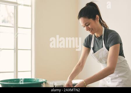 Donna sorridente che indossa grembiule mentre prepara il cibo sul tavolo contro il muro a casa Foto Stock