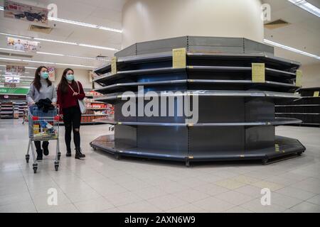 Due donne che indossano le maschere chirurgiche camminano oltre gli scaffali vuoti del supermercato, solitamente immagazzinate con carta igienica e rotoli della cucina.Panic si sviluppa a Hong Kong come la città ha confermato il caso 50th di Coronavirus Nuovo (2019-nCoV). Oltre 40.000 casi di virus hanno confermato in tutto il mondo, ha finora rivendicato oltre 1000 vite. Foto Stock