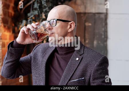 Ricco uomo calvo di mezza età in bicchieri in piedi nel ristorante loft e bere alcolici dal vetro Foto Stock