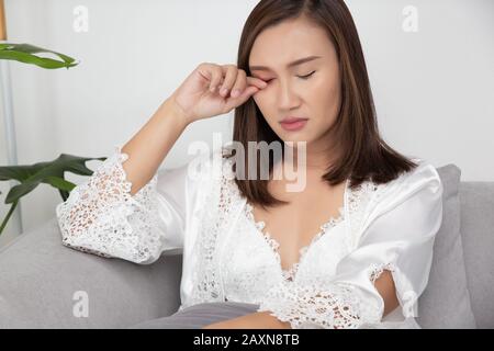 Le donne asiatiche in un abito da notte indossano una veste bianca in raso strofinando l'occhio seduto sul divano grigio nel soggiorno di notte, una ragazza in seta da notte sc Foto Stock