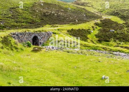 Antica strada che conduce attraverso lussureggianti colline di montagna attraverso un ponte di pietra e un ruscello d'acqua in Highlands scozzesi in estate Foto Stock