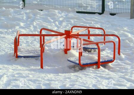 Carosello in metallo rosso per bambini coperto di neve. Concetto di intrattenimento per il tempo libero di strada per i bambini, miglioramento dell'ambiente urbano. Stock foto con spazio vuoto per il testo. Foto Stock