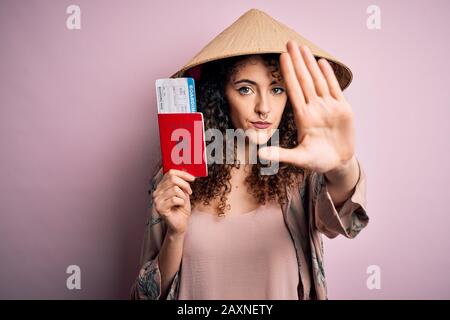 Bella donna turistica con capelli ricci e piercing indossare cappello asiatico tenendo passaporto giapponese con mano aperta facendo segno di stop con serio e confiden Foto Stock