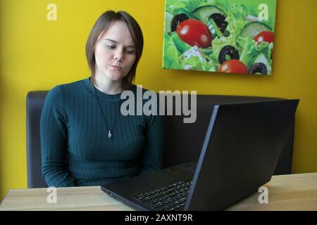 La ragazza con un'espressione scettica pensiva del volto siede su un laptop ad un tavolo in un caffè su un divano marrone contro un muro giallo. Foto Stock