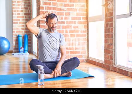 Bell'uomo sportivo di media età seduto sul tappeto facendo esercizio di yoga stretching in palestra confondere e chiedersi circa la domanda. Incerto con dubbio, pensando Foto Stock