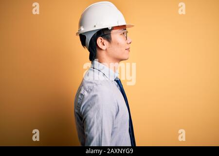 Giovane e bell'architetto cinese uomo che indossa casco di sicurezza e cravatta su sfondo giallo guardando a lato, relax posa profilo con viso naturale con c Foto Stock