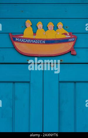 una piccola barca in legno tagliato o modello con quattro persone o una famiglia che indossa impermeabilizzazione in giallo sulla porta di una capanna da spiaggia dipinta in design shabby chic. Foto Stock