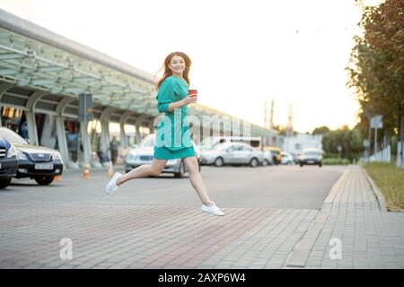 Giovane donna felice corre attraverso il crosswalk. Caffè caldo in un bicchiere di plastica. Abito verde. Il concetto di business, lavoro, stile di vita e tecnologia. Foto Stock