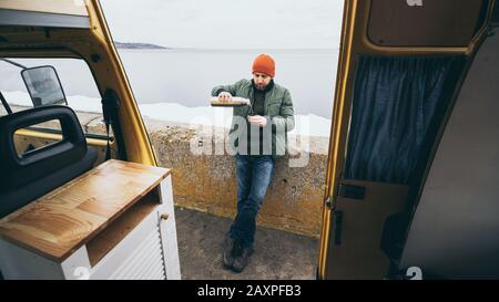 Giovane uomo che beve una bevanda calda dai thermos di fronte al camper che si affaccia su un lago sullo sfondo Foto Stock