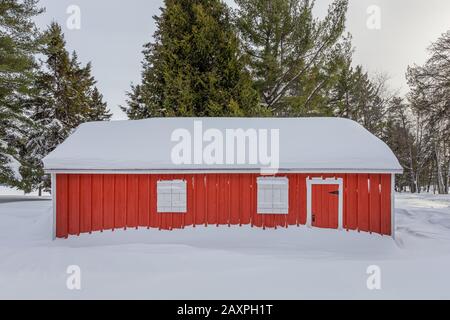 Capannone rosso con persiane bianche incastonato nella neve profonda e circondato da pini verdi. Foto Stock